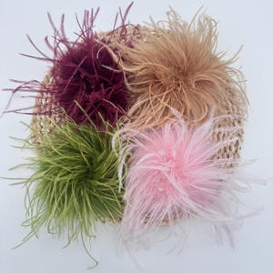 May include: Four round ostrich feather pom-poms in deep burgundy, tan, olive green, and soft pink. Each fluffy, textured plume cluster is arranged on a circular, woven, light-colored natural fiber base, showcasing distinct, wispy feather strands.