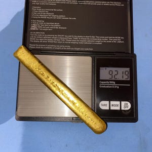 Op de afbeelding: Een lange, getextureerde goudkleurige staaf die op een zwarte digitale weegschaal ligt. De weegschaal heeft een metalen weegplateau en een digitaal display dat het gewicht van 92,19 gram toont, met een capaciteit van 500 gram en 0,01 g.