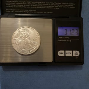 Może przedstawiać: Srebrna moneta American Eagle z 2024 roku spoczywająca na czarnej wadze cyfrowej. Waga posiada metalową platformę ważącą oraz niebieski podświetlany ekran LCD wskazujący 31,23 grama, z przyciskami sterującymi TARE, MODE oraz ON/OFF.