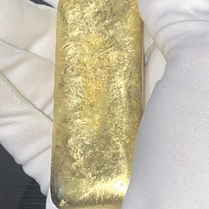 Könnte beinhalten: Ein rechteckiger Goldbarren mit einer texturierten, metallischen Oberfläche. Der Barren hat abgerundete Ecken und ein helles, reflektierendes gelbes Finish. Die Oberfläche weist über die gesamte Länge unregelmäßige, gehämmert wirkende Muster auf.