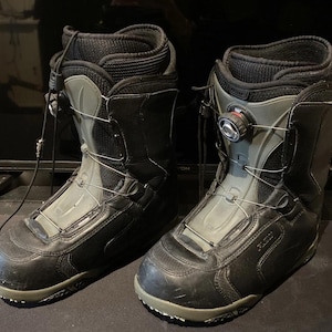Könnte beinhalten: Ein Paar schwarze und graue Snowboardstiefel mit einem BOA-Schnürsystem und einem runden Drehverschluss auf der Vorderseite. Die Stiefel haben ein hohes Design mit gepolsterten Kragen, verstärkten Zehenkappen und einer strukturierten Außenschale.