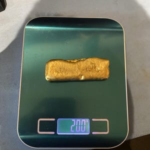 Könnte beinhalten: Ein rechteckiger, goldfarbener Metallbarren mit strukturierter, unebener Oberfläche auf einer dunkelgrünen digitalen Küchenwaage. Das beleuchtete Display der Waage zeigt ein Gewicht von 200 Gramm für das Metallobjekt an.