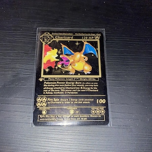 Puede incluir: Una carta coleccionable de Charizard en negro y dorado que muestra al personaje con alas azules escupiendo fuego. La carta muestra 120 HP, el poder Energy Burn y un ataque Fire Spin que inflige 100 de daño. Incluye texto sobre el Flame Pokemon.