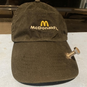 Könnte beinhalten: Eine schwarze Baseballkappe mit dem gelben, aufgestickten McDonald's-Logo auf der Vorderseite. Eine silberfarbene Metallschraube mit Sechskantkopf und eine passende Mutter sind durch den Stoff des Kappen-Schirms auf der rechten Seite befestigt.