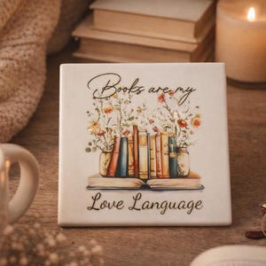 Op de afbeelding: Een vierkante witte keramische tegel met een kleurrijke illustratie van een rij boeken op een open boek, omringd door wilde bloemen. De tekst "Books are my Love Language" is in zwarte letters boven en onder de illustratie gedrukt.