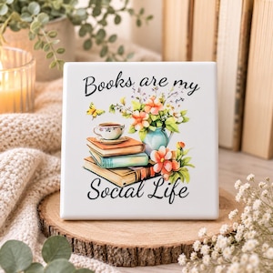 Op de afbeelding: Een vierkante witte keramische tegel met een kleurrijke illustratie van een stapel van drie boeken, een theekopje, een vaas met bloemen en een gele vlinder. De zwarte tekst luidt Books are my Social Life, geplaatst boven en onder de afbeelding.