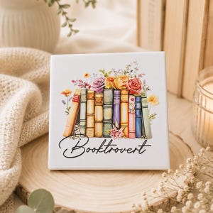 Op de afbeelding: Een vierkante witte keramische tegel met een kleurrijke illustratie van een rij boeken met daarop diverse bloemen, waaronder rozen in roze, oranje en gele tinten. Het woord Booktrovert is in zwarte cursieve letters onder de boeken gedrukt.