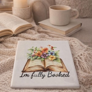 Op de afbeelding: Een vierkante witte keramische tegel met een kleurrijke illustratie van een open boek met een boeket van oranje, paarse, blauwe en gele bloemen die uit de pagina's komen, met de zwarte tekst "I'm fully Booked" onder het boek gedrukt.