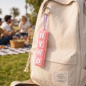 May include: A light pink rectangular keyring featuring four stacked square buttons, each displaying a white letter to spell HYWD. The keyring is attached to a silver metal ring by a purple beaded chain and hangs from a beige canvas backpack.