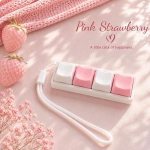 May include: A rectangular white mechanical keyboard macro pad with four cat-ear shaped keycaps in alternating white and pink colours, attached to a white wrist strap. The text Pink Strawberry and A little click of happiness is visible.