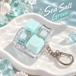 May include: A clear acrylic mechanical keyboard switch keyring featuring two light seafoam green keycaps. The transparent housing reveals the internal switch mechanism. The text Sea Salt Green is printed in the upper right corner.