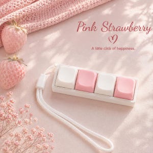 Könnte beinhalten: Ein weißes mechanisches Makro-Tastenfeld mit vier Tasten, die abwechselnd weiß und rosa sind. Das Gerät verfügt über eine weiße Handschlaufe und ist mit dem Text Pink Strawberry und A little click of happiness bedruckt.