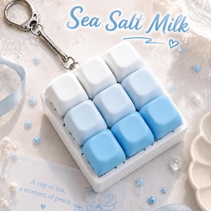 May include: A square keychain featuring a grid of nine smooth, matte keycaps in a gradient of white to light blue. The text Sea Salt Milk is printed above the keychain, and a small card below reads A cup of tea, a moment of peace.