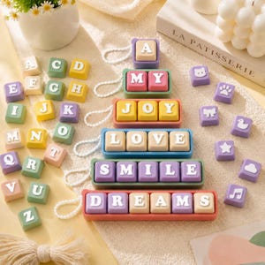 May include: A collection of colourful square keycap-style ornaments featuring white letters and symbols. The pieces are arranged in a pyramid shape with words like MY, JOY, LOVE, SMILE, and DREAMS, accompanied by loose alphabet and icon keys.
