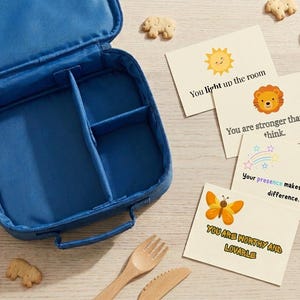 Pode incluir: Uma lancheira azul com zíper e divisórias internas, acompanhada por quatro cartões retangulares com ilustrações de um sol, leão, arco-íris e borboleta, junto com textos como You light up the room e You are stronger than you think.