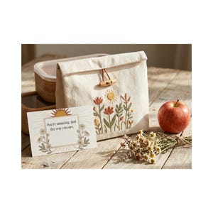 May include: A beige canvas lunch bag with an embroidered floral design, a wooden toggle closure, and a greeting card that reads "You're amazing, just the way you are." An apple and a wooden container are also present.