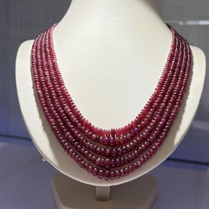 Op de afbeelding: Een meerrijige ketting bestaande uit vijf rijen kleine, ronde, dieprode kralen. De kralen zijn uniform van vorm en grootte, wat zorgt voor een gelaagd, trapsgewijs effect rond de hals. De dieprode kleur is consistent over alle vijf de strengen.