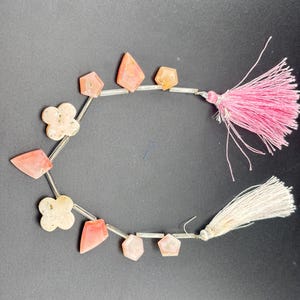 May include: A strand of assorted peach-coloured stone beads in geometric and flower shapes, interspersed with clear cylindrical spacer beads. The strand is finished with one bright pink tassel and one off-white tassel at the ends.