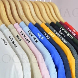 Puede incluir: Una fila de camisetas colgadas en perchas de madera en varios colores, incluyendo blanco, gris ceniza, arena, rosa claro, azul claro, real, marino, oro, gris oscuro jaspeado, negro, verde bosque, granate y rojo, con cada color etiquetado.
