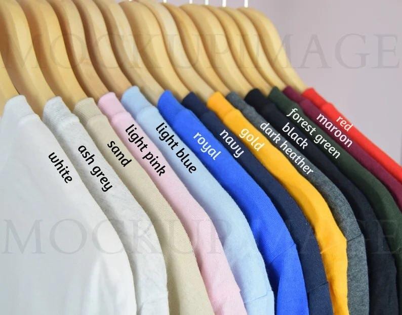 Puede incluir: Una fila de camisetas colgadas en perchas de madera en varios colores, incluyendo blanco, gris ceniza, arena, rosa claro, azul claro, real, marino, oro, gris oscuro jaspeado, negro, verde bosque, granate y rojo, con cada color etiquetado.