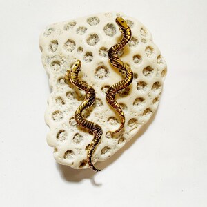 May include: A pair of gold-toned snake-shaped earrings featuring a textured, segmented body design with a curved, serpentine silhouette. Each earring displays a detailed head and a tapering tail, showcasing a polished metallic finish throughout.