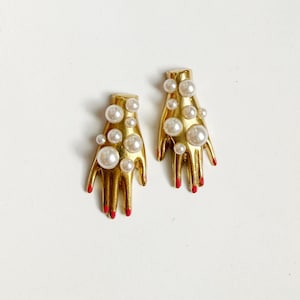 Pode incluir: Um par de brincos dourados em forma de mão. Cada brinco apresenta uma mão com unhas pintadas de vermelho e é adornado com várias pérolas falsas de vários tamanhos. Os brincos estão sobre um fundo branco.