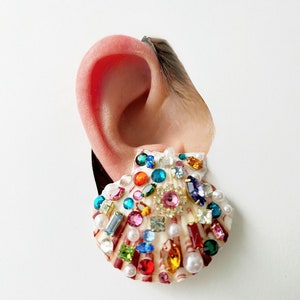 May include: A seashell earring adorned with an array of colorful gemstones and pearls. The earring features a variety of shapes and sizes of gems, including round, square, and rectangular, in shades of pink, blue, green, and orange.