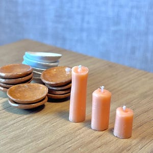 May include: A collection of miniature dollhouse items. There are stacks of small brown plates, white and blue plates, and three peach-coloured candles of different heights. The items are on a wooden surface.