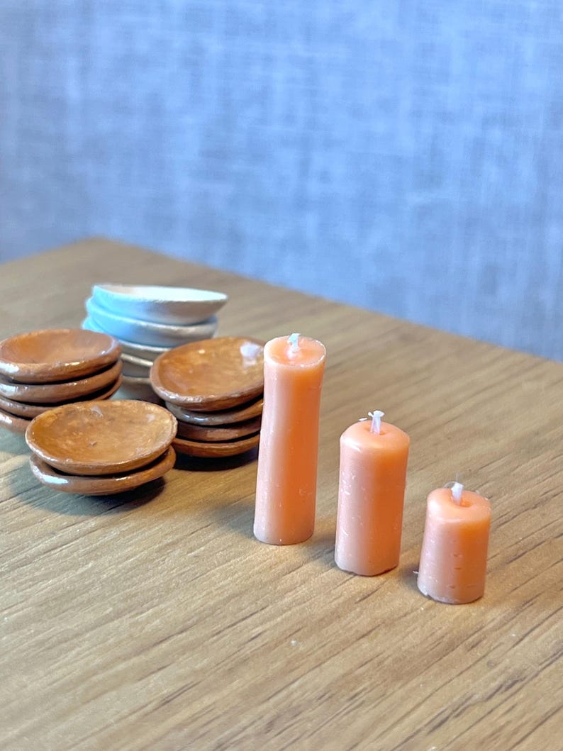 May include: A collection of miniature dollhouse items. There are stacks of small brown plates, white and blue plates, and three peach-coloured candles of different heights. The items are on a wooden surface.