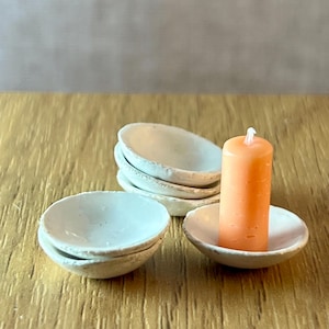 May include: A set of small, off-white ceramic bowls and an orange candle. The candle sits in a matching bowl. The bowls are stacked and arranged on a wooden surface, creating a minimalist aesthetic.
