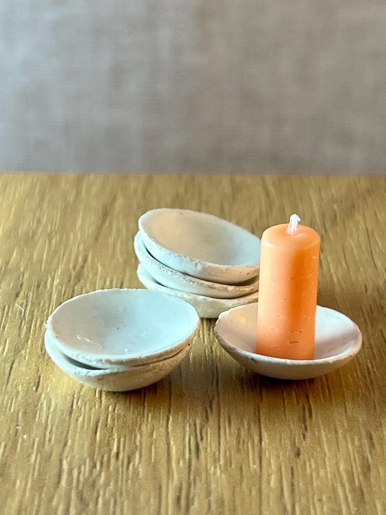 May include: A set of small, off-white ceramic bowls and an orange candle. The candle sits in a matching bowl. The bowls are stacked and arranged on a wooden surface, creating a minimalist aesthetic.