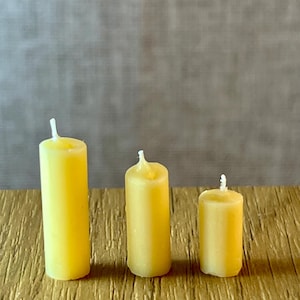 May include: Three yellow pillar candles of different heights. The candles are cylindrical with a white wick. They are arranged on a textured, gold-coloured surface, with a neutral grey background.