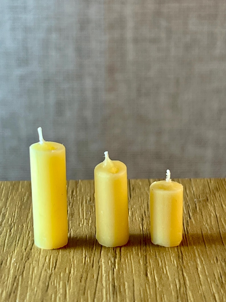 May include: Three yellow pillar candles of different heights. The candles are cylindrical with a white wick. They are arranged on a textured, gold-coloured surface, with a neutral grey background.
