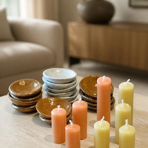 May include: A display of decorative items featuring stacks of small, brown and white ceramic dishes and pillar candles in shades of orange and yellow. The candles vary in height and are arranged on a neutral surface.