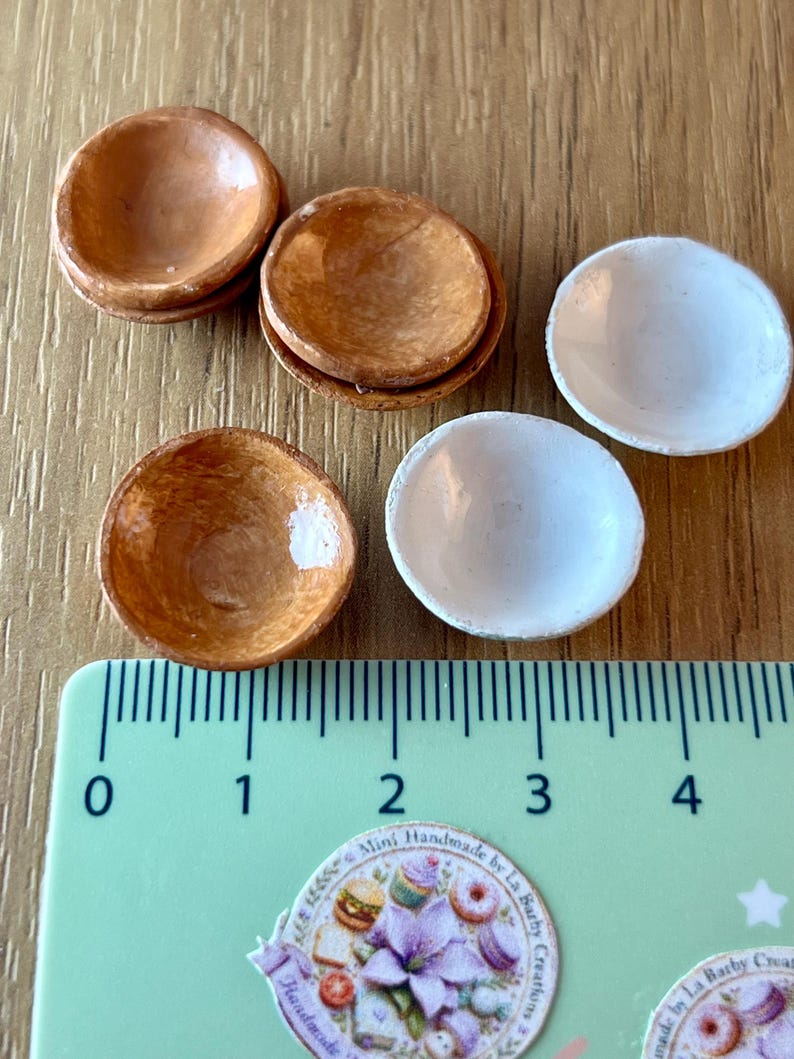 May include: Miniature wooden bowls in shades of brown and white. The bowls are arranged on a wooden surface next to a ruler. The ruler shows measurements in centimetres. The bowls are handmade.