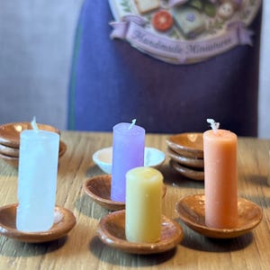 May include: Miniature candles in white, purple, yellow, and orange, each on a small, round, brown dish. The background features a purple apron with the text "Handmade Miniatures". The candles are likely for decorative purposes.