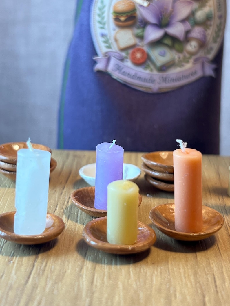May include: Miniature candles in white, purple, yellow, and orange, each on a small, round, brown dish. The background features a purple apron with the text "Handmade Miniatures". The candles are likely for decorative purposes.