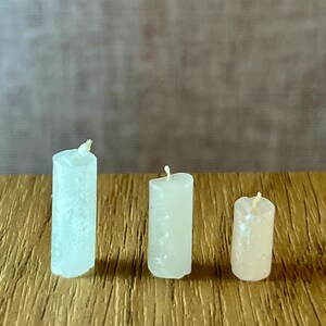 May include: Three small, cylindrical candles of varying heights and colours are arranged on a wooden surface. The candles range in colour from white to pale yellow, each with a visible wick at the top. The wooden surface has a textured, golden-brown appearance.