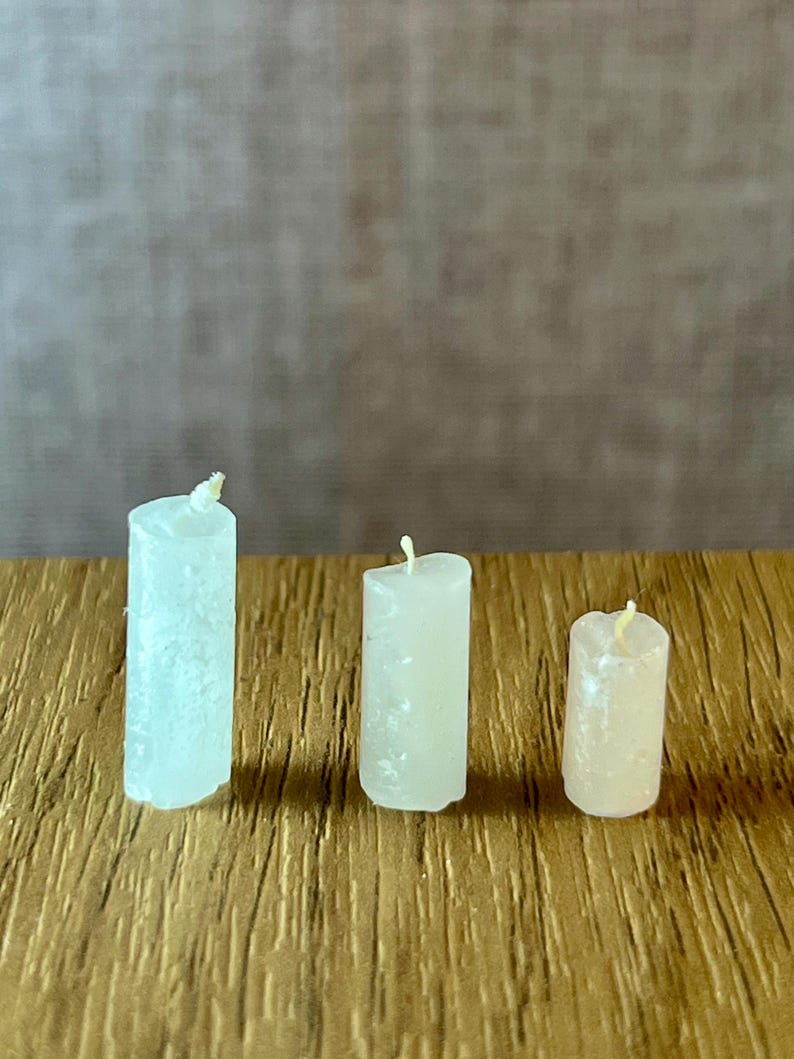 May include: Three small, cylindrical candles of varying heights and colours are arranged on a wooden surface. The candles range in colour from white to pale yellow, each with a visible wick at the top. The wooden surface has a textured, golden-brown appearance.