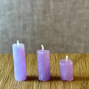 May include: Three small, lavender pillar candles of different heights are displayed on a wooden surface. Each candle has a white wick. The candles have a textured surface. The candles are in centimetres.