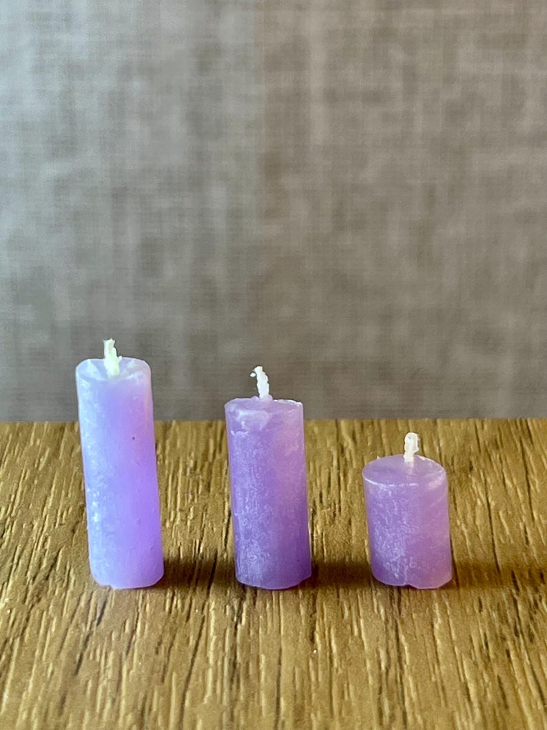 May include: Three small, lavender pillar candles of different heights are displayed on a wooden surface. Each candle has a white wick. The candles have a textured surface. The candles are in centimetres.