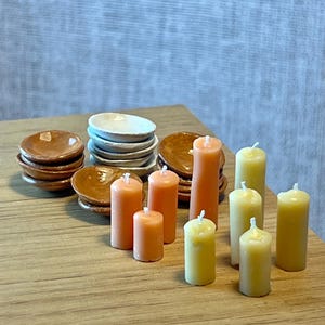 May include: A collection of miniature dollhouse items, including stacks of small plates in brown and white, and several orange and yellow pillar candles. The items are arranged on a wooden surface, suggesting a dining or decorative theme.