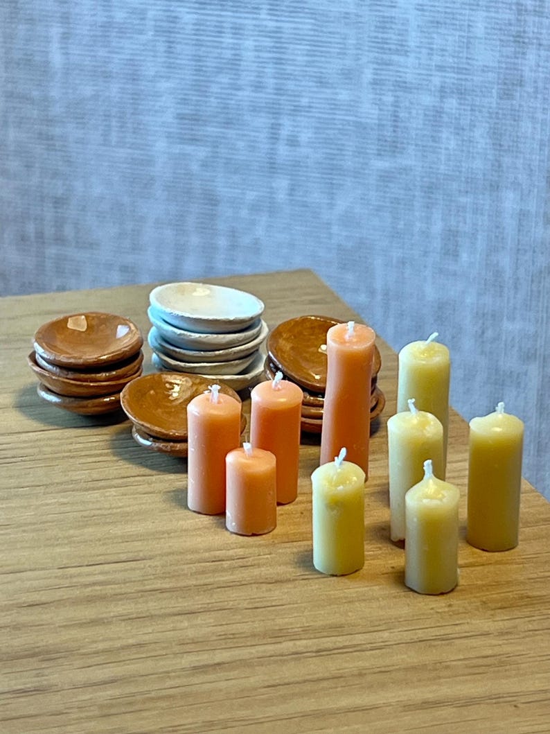 May include: A collection of miniature dollhouse items, including stacks of small plates in brown and white, and several orange and yellow pillar candles. The items are arranged on a wooden surface, suggesting a dining or decorative theme.