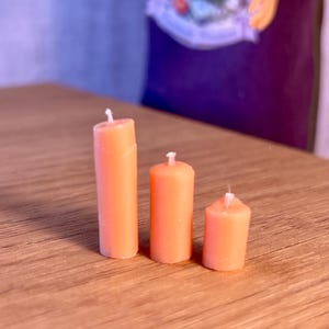 May include: Three orange pillar candles of different heights are displayed on a wooden surface. Each candle has a white wick. The candles are cylindrical and a solid orange colour.