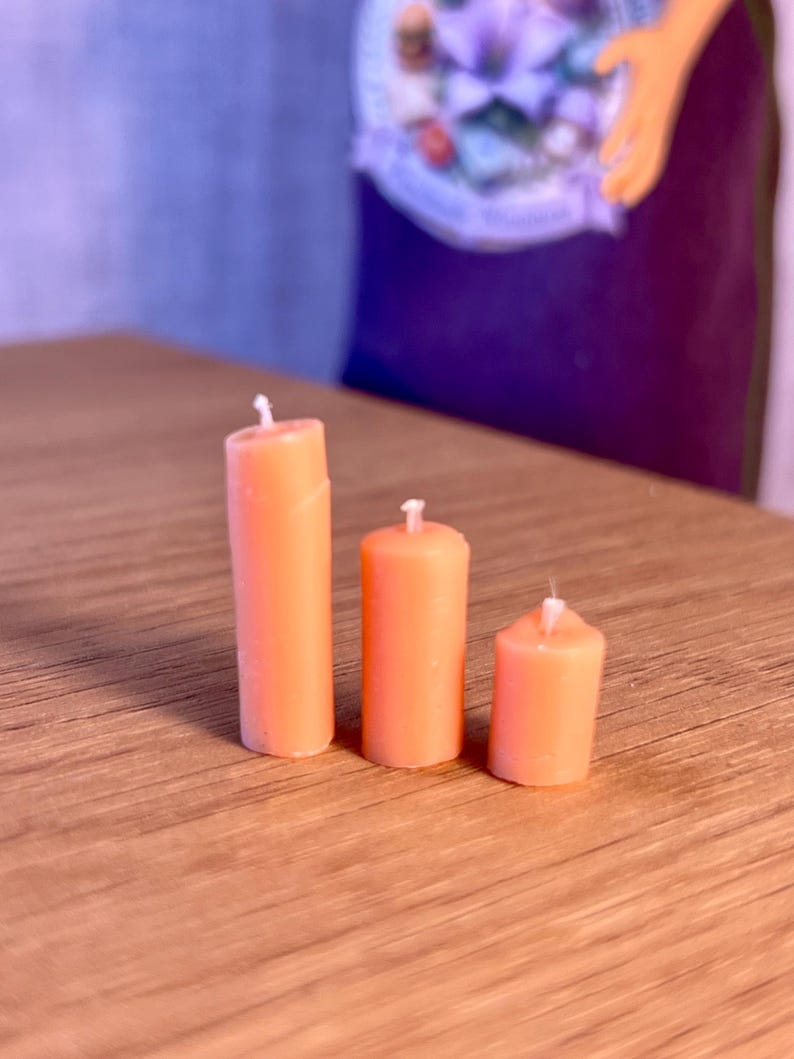 May include: Three orange pillar candles of different heights are displayed on a wooden surface. Each candle has a white wick. The candles are cylindrical and a solid orange colour.