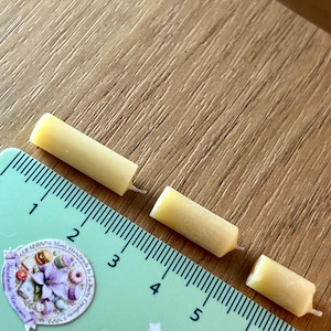 May include: Three small, yellow candles are positioned near a ruler. The candles are cylindrical with a wick. The ruler is green with black markings and the text "Mini Handmade by La Barby Creations". The candles are approximately 1.5 cm long.