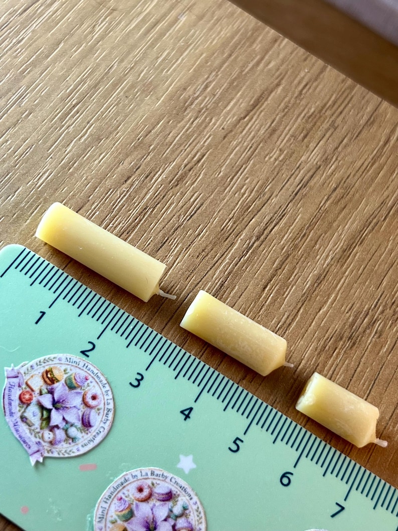 May include: Three small, yellow candles are positioned near a ruler. The candles are cylindrical with a wick. The ruler is green with black markings and the text "Mini Handmade by La Barby Creations". The candles are approximately 1.5 cm long.