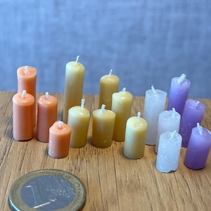 May include: A selection of miniature pillar candles in various colours, including orange, yellow, white, and purple. The candles are displayed on a wooden surface, with a Euro coin for scale. Each candle has a white wick.