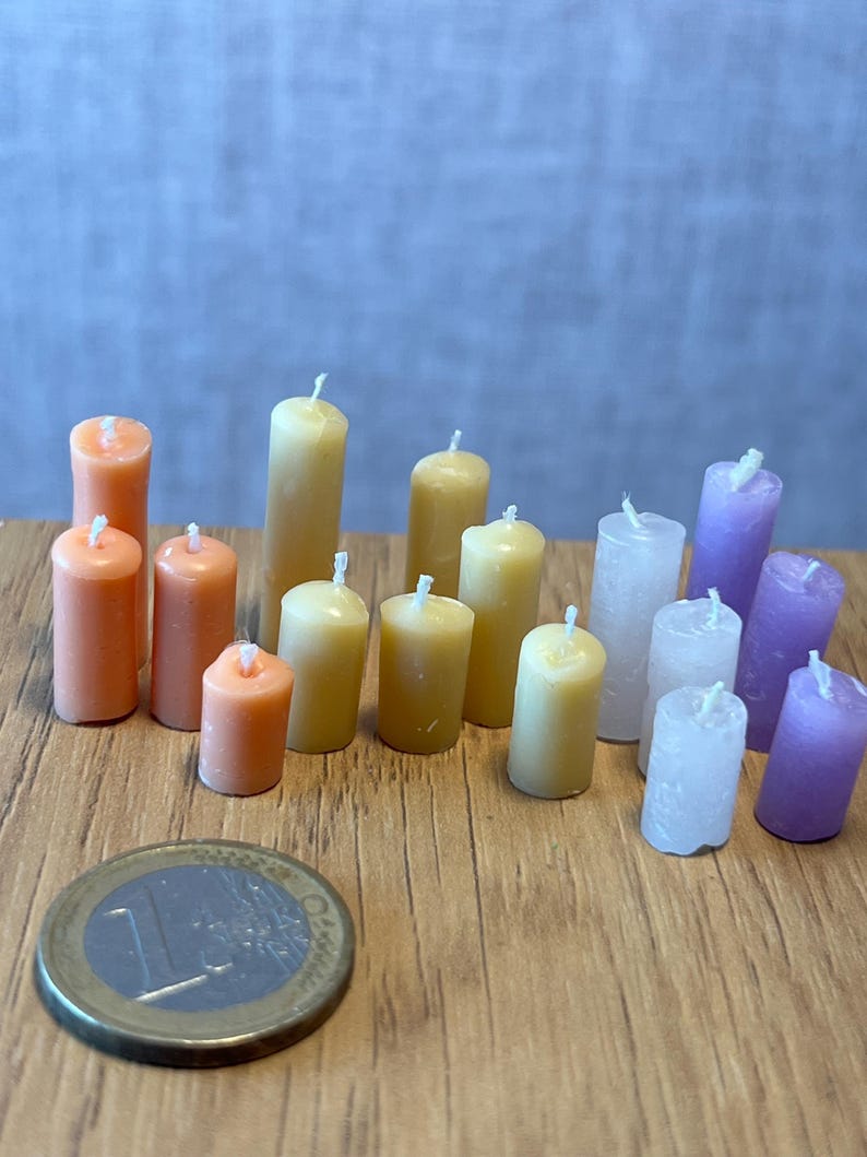 May include: A selection of miniature pillar candles in various colours, including orange, yellow, white, and purple. The candles are displayed on a wooden surface, with a Euro coin for scale. Each candle has a white wick.