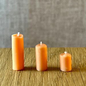 May include: Three orange pillar candles of different heights are displayed on a textured, golden-brown surface. Each candle has a white wick. The background is a neutral grey. The candles are measured in centimetres.
